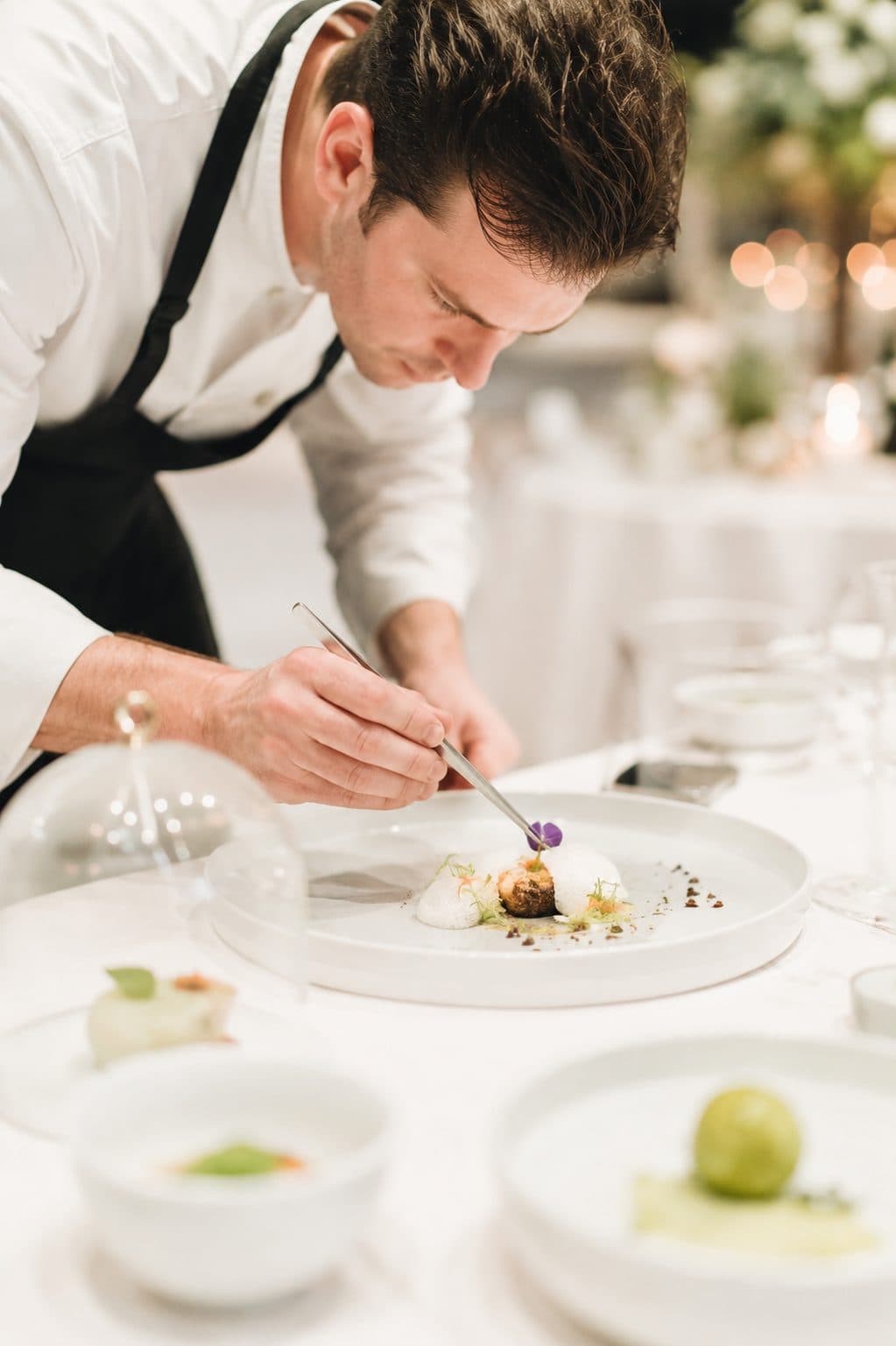 Chef plating a dish at an event table, candlelight behind