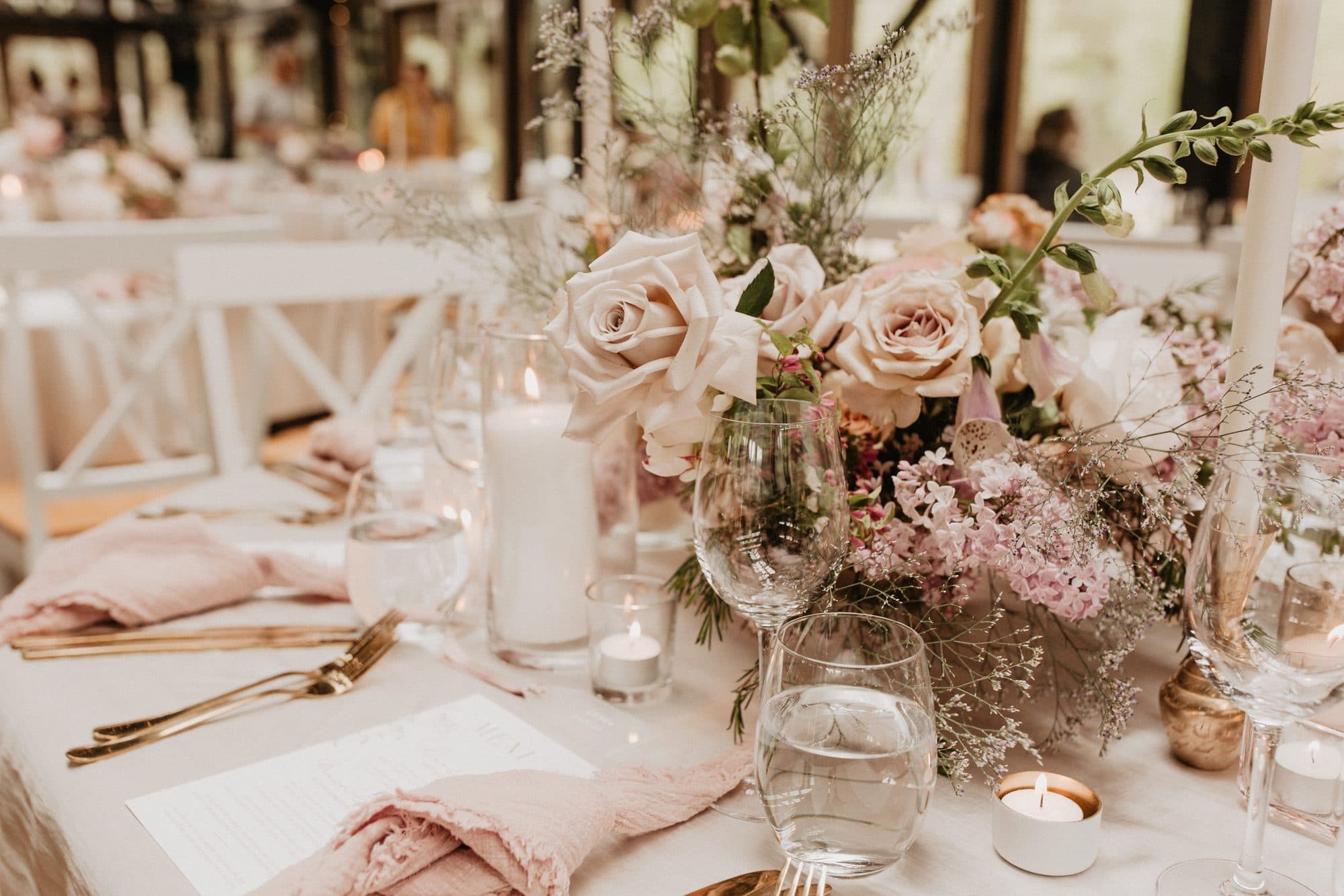 Long table dressed in blush linen with roses, pillar candles and gold cutlery